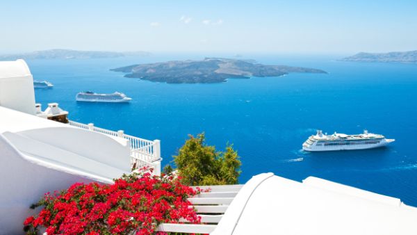 Cruise ships anchored in the blue waters of Santorini's caldera, viewed from a whitewashed hillside with colorful bougainvillea