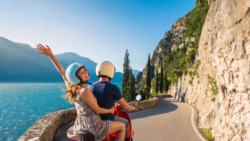 Deux personnes conduisant un scooter rouge sur une route panoramique le long d'un grand plan d'eau entouré de montagnes