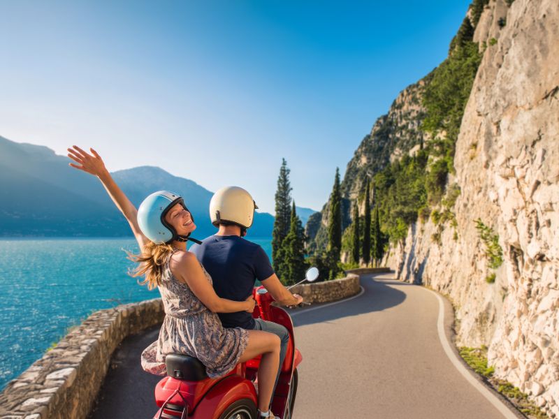 Two people riding a red scooter on a scenic road along a large body of water surrounded by mountains
